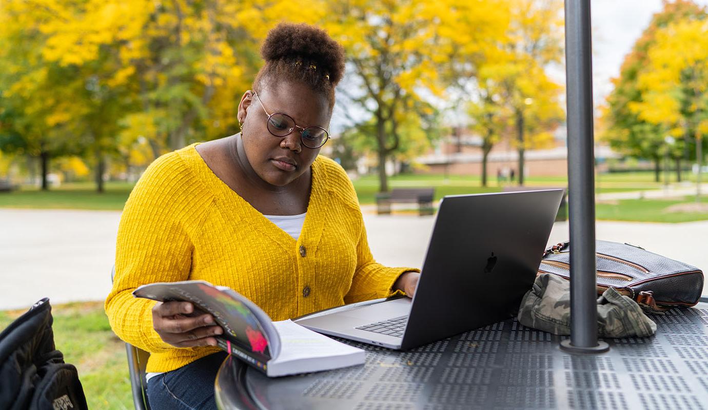 Student sitting outside at table with laptop and book